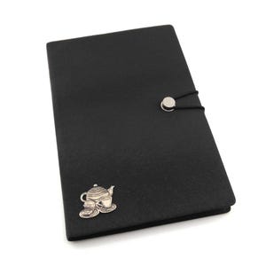May include: Black leather-effect journal featuring a silver teapot and teacup emblem. The journal has a button closure with a black elastic cord. The cover is plain and smooth, with a subtle texture.