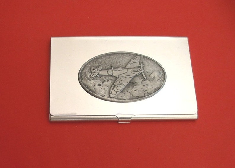 Puede incluir: Un porta tarjetas de visita de metal plateado con un dise&ntilde;o en relieve de forma ovalada que representa un avi&oacute;n de combate de la Segunda Guerra Mundial en vuelo. El avi&oacute;n est&aacute; representado en colores de camuflaje y vuela sobre las nubes.