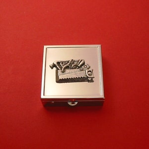 May include: A silver metal pill box with a raised image of a toolbox filled with tools. The toolbox is silver and the tools are a variety of shapes and sizes.