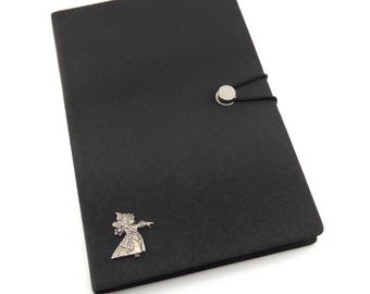 May include: Black leather journal with a silver metal clasp and a decorative figure. The journal has a button closure with a black elastic cord. The figure is a stylized character in a costume.