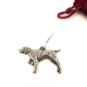 Może przedstawiać: Srebrny charm w kształcie psa, stojącego na czterech łapach z głową zwróconą w bok. Charm jest przymocowany do srebrnej pętli.
