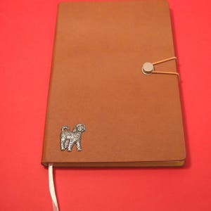 Può includere: Un diario in pelle marrone con una silhouette di cane argentata sulla copertina. Il diario ha una chiusura a bottone argentata e un segnalibro a nastro bianco.
