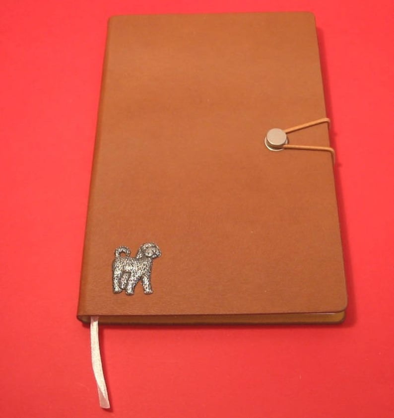 K&ouml;nnte beinhalten: Ein braunes Lederjournal mit einer silbernen Hundesilhouette auf dem vorderen Umschlag. Das Journal hat einen silbernen Knopfverschluss und ein wei&szlig;es Lesezeichenband.