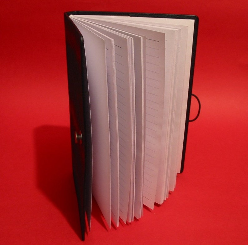 K&ouml;nnte beinhalten: Schwarzes Notizbuch mit linierten wei&szlig;en Seiten, ge&ouml;ffnet, um das Innere zu zeigen. Das Notizbuch hat einen schwarzen elastischen Verschluss und einen Knopfverschluss. Die Seiten sind aufgef&auml;chert und zeigen das linierte Papier. Der Hintergrund ist einfarbig rot.