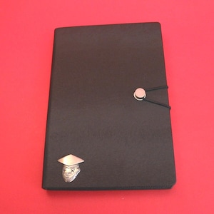 Op de afbeelding: Een zwart rechthoekig notitieboek met een textuur op de kaft, vastgezet met een zwarte elastische band aan een zilveren ronde knop. Een klein zilveren embleem van een gezicht met een afstudeerhoed bevindt zich in de linkerbenedenhoek.