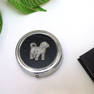 Peut inclure: Une boîte à pilules ronde argentée avec un centre en similicuir noir. Une figurine de chien détaillée de couleur argentée est au centre. La boîte à pilules est sur une surface blanche, avec des feuilles vertes en arrière-plan.