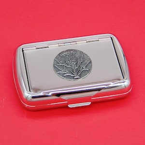 May include: A silver metal cigarette case with a hinged lid. The case features an embossed design of a thistle flower.