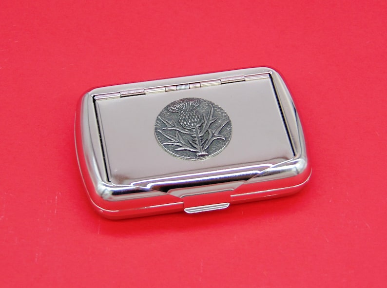 May include: A silver metal cigarette case with a hinged lid. The case features an embossed design of a thistle flower.