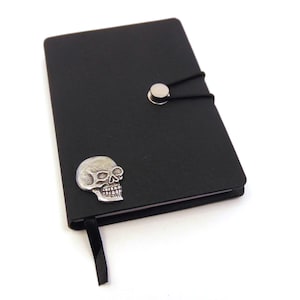 Puede incluir: Diario de cuero negro con un diseño de calavera plateada y un cierre elástico negro con un botón plateado.