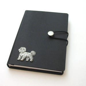 Puede incluir: Cuaderno negro con un charm de perro Bichon Frise plateado. El cuaderno tiene un cierre de botón con una banda elástica negra. El charm de perro es detallado y brillante. El cuaderno está sobre un fondo blanco.