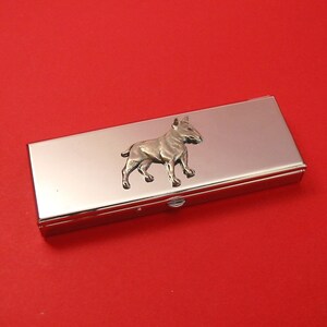 May include: A silver pill box with a raised image of a white bull terrier dog on the lid.