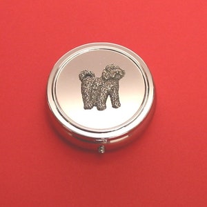 May include: A silver round pill box with a small, detailed image of a white dog on the lid.