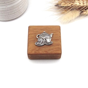 May include: A small square wooden box featuring a silver-toned metal emblem on the lid. The emblem depicts a teapot, a teacup on a saucer, and a small cupcake on a plate. The wood has a natural grain pattern and a smooth, polished finish.