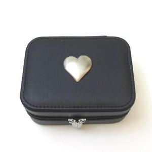 Peut inclure: Une boîte à bijoux noire et rectangulaire avec un emblème argenté en forme de cœur sur le dessus. La boîte a une fermeture éclair et est probablement en cuir ou dans un matériau similaire. Le fond est blanc.
