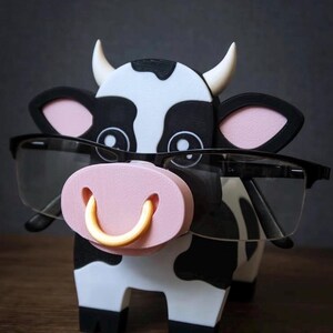 May include: A whimsical cow-shaped eyeglasses holder. The holder is black and white with a pink nose and a gold-colored ring. The cow is wearing black-framed glasses. The holder is designed to hold eyeglasses.