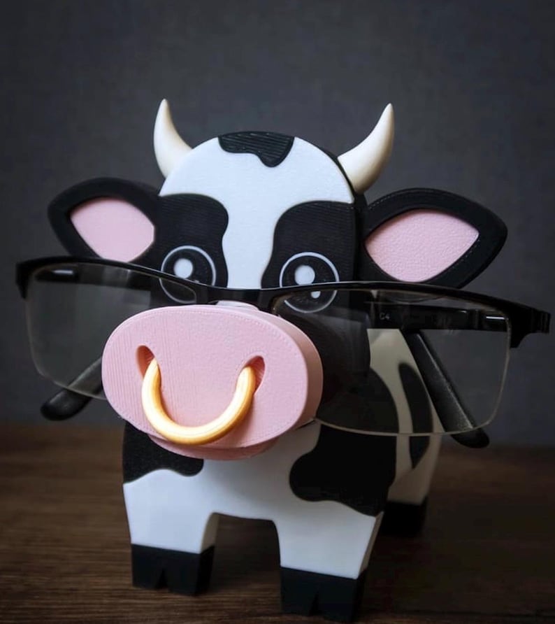 May include: A whimsical cow-shaped eyeglasses holder. The holder is black and white with a pink nose and a gold-colored ring. The cow is wearing black-framed glasses. The holder is designed to hold eyeglasses.
