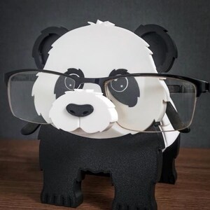 May include: A black and white panda-shaped eyeglasses holder. The holder is designed to look like a panda with black ears, eyes, and body, and a white face and chest. The panda is wearing a pair of black-framed glasses.