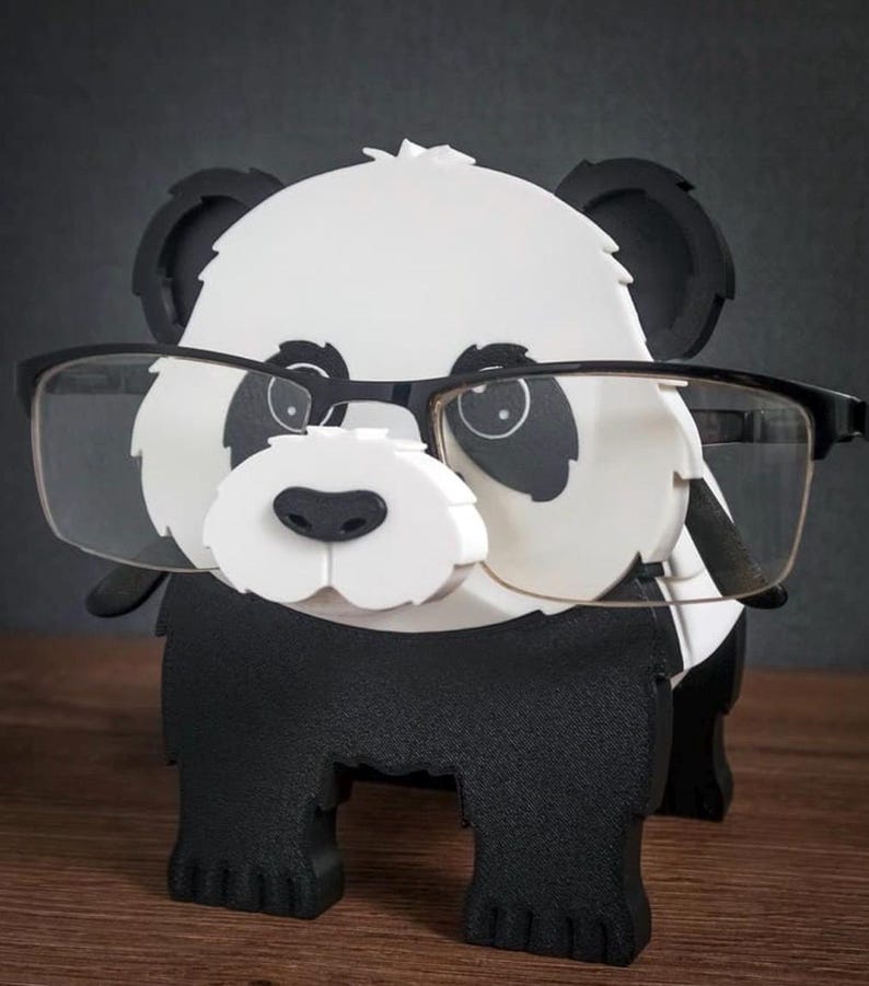 May include: A black and white panda-shaped eyeglasses holder. The holder is designed to look like a panda with black ears, eyes, and body, and a white face and chest. The panda is wearing a pair of black-framed glasses.