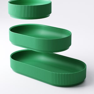May include: Three green, oval-shaped serving trays stacked in a vertical composition. The trays have a ribbed exterior and a smooth interior. The trays are a vibrant green color and are set against a white background.
