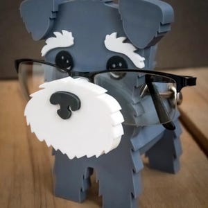 May include: A gray dog figurine with white accents and black glasses. The dog has a white beard and eyebrows, and black button eyes. The glasses are black-framed and sit on the dog's nose. The figurine is standing.
