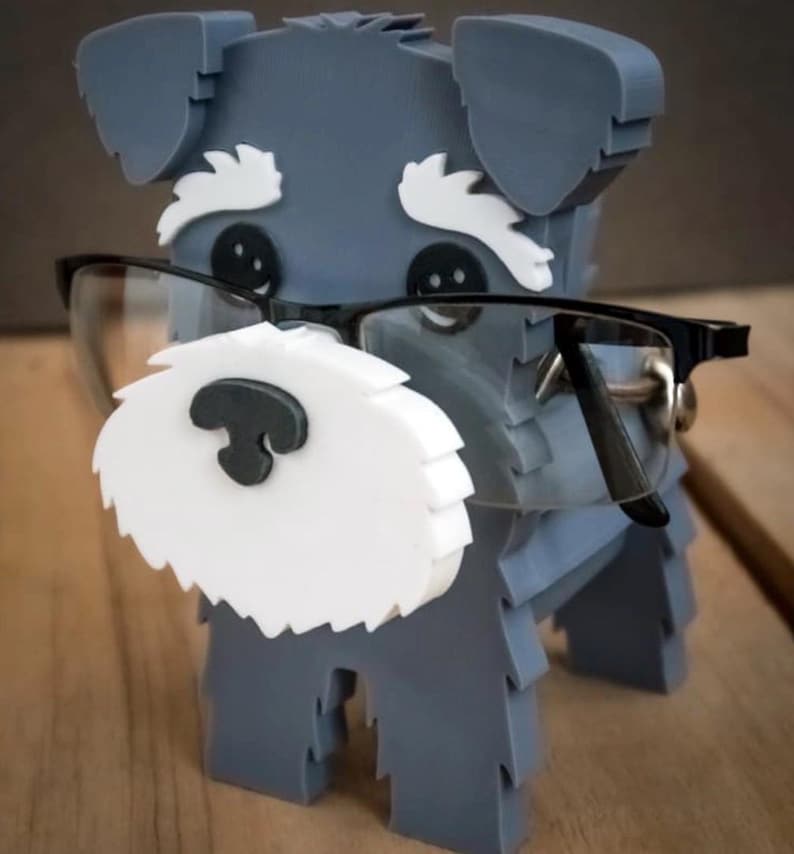 May include: A gray dog figurine with white accents and black glasses. The dog has a white beard and eyebrows, and black button eyes. The glasses are black-framed and sit on the dog's nose. The figurine is standing.