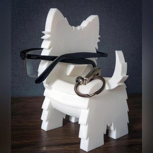 May include: A white, dog-shaped eyeglasses holder with black-framed glasses resting on the head. A silver key ring is attached to the dog's body. The holder is made of a solid, textured material.