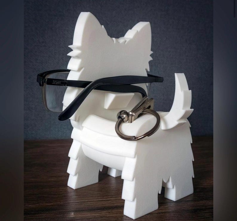 May include: A white, dog-shaped eyeglasses holder with black-framed glasses resting on the head. A silver key ring is attached to the dog's body. The holder is made of a solid, textured material.
