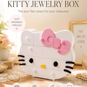 May include: A white cat-shaped jewelry box with a pink bow, black whiskers, black eyes, and a yellow nose. The front features multiple drawers with small white knobs. Text on the box includes "Sweet. Simple. So You. KITTY JEWELRY BOX".