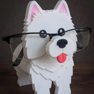 May include: A white dog-shaped eyeglass holder with black-rimmed glasses. The dog has pink inner ears, a black nose, and a red tongue. The holder is made of a textured material and sits on a wooden surface.