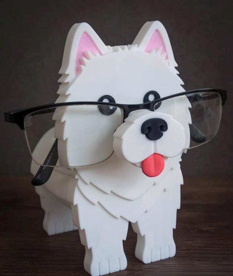 May include: A white dog-shaped eyeglass holder with black-rimmed glasses. The dog has pink inner ears, a black nose, and a red tongue. The holder is made of a textured material and sits on a wooden surface.