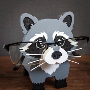 May include: A gray and white raccoon-shaped glasses holder with black accents. The holder has a 3D design with a black nose, white whiskers, and black-framed glasses resting on its face. The product is made of wood.