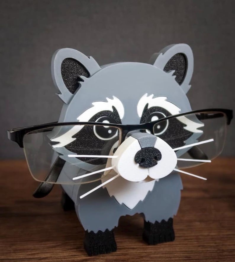 May include: A gray and white raccoon-shaped glasses holder with black accents. The holder has a 3D design with a black nose, white whiskers, and black-framed glasses resting on its face. The product is made of wood.
