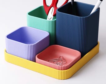 Könnte beinhalten: Ein Schreibtisch-Organizer mit mehreren Fächern, bestehend aus einer gelben Basis, einem hohen dunkelblauen Becher, einem mintgrünen Becher, einem lavendelfarbenen Becher und einer kleinen korallenfarbenen Schale. Rote Schere, Stift und Kugelschreiber.