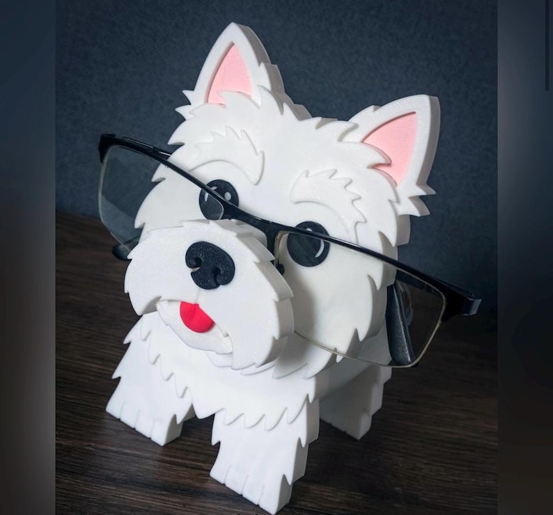 May include: A white dog-shaped eyeglass holder with black-rimmed glasses. The dog has pink inner ears, a black nose, and a red tongue. The holder is made of a textured material and sits on a wooden surface.