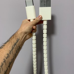 May include: A white USB-C to USB-A cable with a unique design. The cable features a series of white, cube-shaped links connecting the connectors. The connectors are white with silver-colored metal inserts. The cable is held against a gray background.