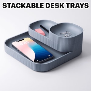 May include: Stackable desk trays in a light blue-grey color. The trays have a ribbed texture and are arranged in a tiered configuration. A smartphone, pencil, eraser, and paper clips are visible. The text "STACKABLE DESK TRAYS" is at the top.