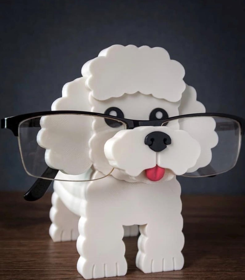 May include: A white dog-shaped eyeglasses holder with black-rimmed glasses. The dog has a pink tongue and black eyes and nose. The holder is made of a solid material and is designed to hold eyeglasses.