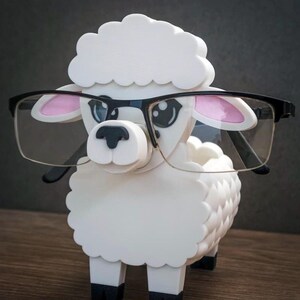 May include: A white sheep figurine wearing black-framed glasses. The sheep has pink inner ears and black hooves. The figurine is made of a smooth, solid material and is posed on a wooden surface.
