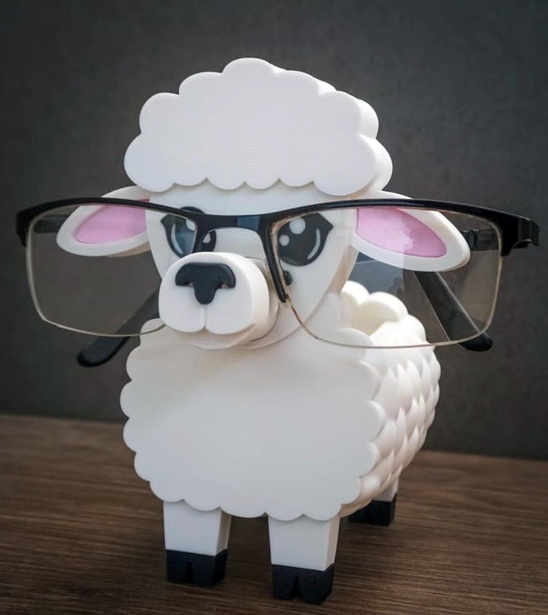 May include: A white sheep figurine wearing black-framed glasses. The sheep has pink inner ears and black hooves. The figurine is made of a smooth, solid material and is posed on a wooden surface.