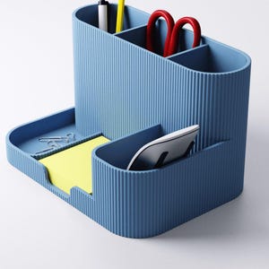 May include: A blue desk organizer with multiple compartments. It holds a pen, pencil, red scissors, sticky notes, paper clips, and a computer mouse. The organizer has a ribbed texture and a modern design.
