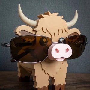 May include: A whimsical, light brown Highland cow-shaped glasses holder with a pink nose and cream-colored horns. The cow is wearing sunglasses with dark lenses. The holder is made of wood and has a textured appearance.