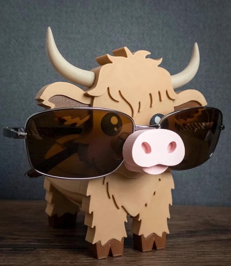 May include: A whimsical, light brown Highland cow-shaped glasses holder with a pink nose and cream-colored horns. The cow is wearing sunglasses with dark lenses. The holder is made of wood and has a textured appearance.