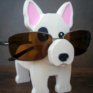 May include: A white dog-shaped eyeglass holder with pink inner ears and a black nose. The holder is wearing brown-tinted sunglasses. The dog has a simple, cartoonish design and is standing on a wooden surface.