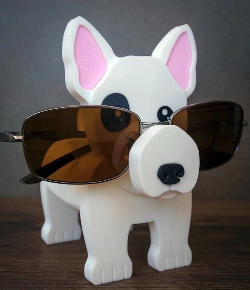 May include: A white dog-shaped eyeglass holder with pink inner ears and a black nose. The holder is wearing brown-tinted sunglasses. The dog has a simple, cartoonish design and is standing on a wooden surface.