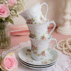 Peut inclure: Trois tasses à thé en porcelaine blanche empilées verticalement sur deux soucoupes assorties. Chaque tasse et soucoupe présente une texture délicatement gaufrée et un motif floral coloré composé de petites roses roses et de feuilles vertes.