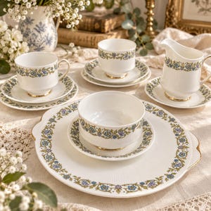 Może przedstawiać: Zestaw do herbaty z białej porcelany z delikatnym wzorem niebiesko-zielonych kwiatowych pnączy i złotym wykończeniem. Komplet zawiera duży półmisek, dzbanek na śmietankę, miseczkę oraz filiżanki ze spodkami, wszystkie o delikatnie żebrowanej fakturze.