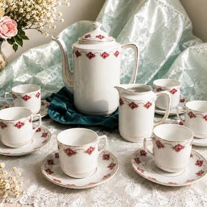 Pode incluir: Um conjunto de chá em porcelana branca composto por um bule alto, uma leiteira e cinco chávenas com pires a condizer. Cada peça é decorada com um padrão delicado de pequenas rosas vermelhas e folhas verdes ligadas por finas linhas douradas.