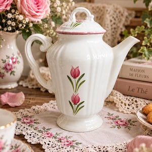 Op de afbeelding: Een witte keramische theepot met een geribbelde behuizing, voorzien van een roze tulpenmotief op de voorzijde en het deksel. De theepot heeft een gebogen handvat, een puntige tuit en een bijpassend deksel met een decoratieve knop.