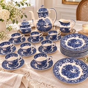 Może przedstawiać: Ceramiczny zestaw do herbaty z niebiesko-białym wzorem przedstawiającym krajobrazy i kwiatowe obwódki. Zestaw zawiera dzbanek do herbaty, cukiernicę z pokrywką, dzbanek na mleko, sześć filiżanek ze spodkami oraz stos sześciu talerzy obiadowych.