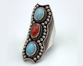 May include: A silver ring with three oval-shaped stones. The top and bottom stones are light blue, and the center stone is red. The ring has a dark brown border and a beaded design around the stones.